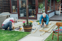Adult Son And Father Carpenter Working Together, Handle Wooden Timbers In The Garden. Patio Construction By Your Own. DIY, Do It Yourself. Home Renovation, Improvement, Refurbishment. Selective Focus