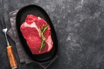 Wall Mural - Beef steak. Fresh raw beef rib eye steak with fork rosemary, salt and pepper on a grill pan on dark grey or black stone background. Shop advertising idea. Top view. Mockup for design idea.