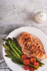 Wall Mural - Chicken breast grilled with vegetables. Grilled chicken steak, asparagus and cherry tomatoes in black plate on light gray concrete table background. Healthy diet lunch. Barbecue steak fried. Top view.