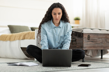 Poster - Serious young woman using laptop, having remote job, sitting on floor in bedroom, full length