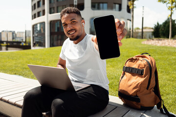 Poster - Happy black guy with laptop showing smartphone with blank screen, recommending mobile app or website, mockup