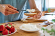 Woman making summer strawberry sandwich. Female hands spread stracchino cheese on bread for toast. Healthy eating, fruit dieting brunch.