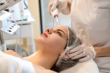 Wall Mural - Beautician applying electric Darsonval facial massage treatment with special tool over patients face in beauty salon