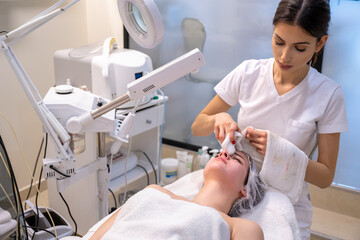 Wall Mural - Female therapist cleaning young woman face after skin treatment in beauty salon.