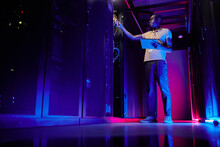 Full Length Portrait Of African American System Administrator Setting Up Server Network In Data Center Lit By Neon Light, Copy Space