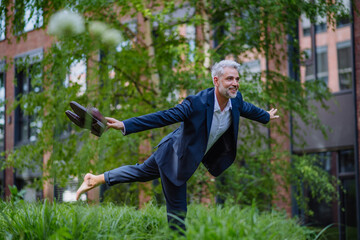 Fun portrait of happy energetic mature businessman carrying shoes and walking barefoot and making pose in park, feeling free, work life balance concept.