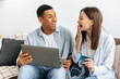 © Kateryna - Excited happy stylish multiracial couple in love, spouses, sitting on sofa in a living room, using laptop and credit card for online shopping, delivery, looking amazed at each other, smiling