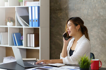 Wall Mural - Happy young Asian business woman sitting smiling and talking on mobile phone in office.