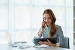 © amnaj - Happy young Asian business woman sitting smiling and talking on mobile phone in office.