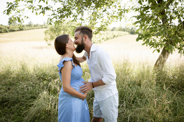 Wall Mural - young couple in love standing in high flower meadow in summer and cuddling and the woman is pregnant and has a blue dress on and the man has a full beard