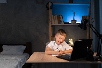 boy is doing homework using a laptop computer at his bedroom desk at night