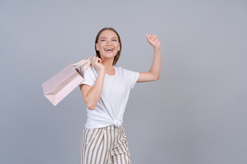 Wall Mural - View lovely slim cheerful girl holding package bag, dancing with pleasure, isolated on gray pastel color background in light clothes. Beautiful young woman rejoices in shopping, mock up and copy space