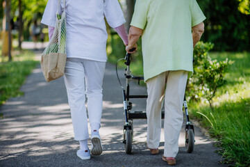 Rear view of caregiver with senior woman lowsection on walk with walker in park with shopping bag.