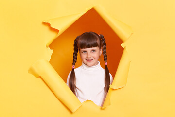 Portrait of happy satisfied little girl with braids wearing white turtleneck posing in torn hole of yellow paper wall, looking at camera with positive optimistic facial expression.