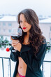 Portrait of young woman wearing unbuttoned black shirt, red underpants standing on balcony, holding glass with red wine.