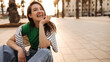 © Look! - Happy young caucasian girl laughs looking away, sits on pavement in middle of square on street. Brunette wears T-shirt, jeans and jacket. Mood, lifestyle, concept.
