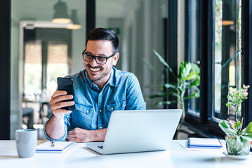 Smiling freelancer with laptop text messaging on smart phone at home office