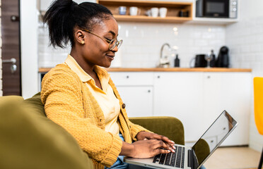 Wall Mural - Young African American woman sitting on sofa at home studying using laptop.	