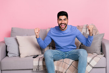 Poster - Photo of funny delighted handsome guy sit couch raise fists celebrate success triumph isolated on pink color background