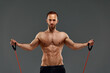 A topless sportsman performs fitness exercises with rubber band in a studio on gray background. Body-building. Athlete.