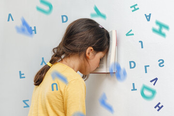 Sad and tired caucasian girl with dyslexia holds a book on her forehead. Flying tangled letters in the air. The child learns to speak and read correctly