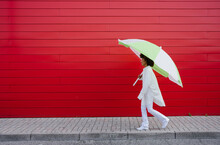 Young Woman Walking With Umbrella On Footpath By Red Wall
