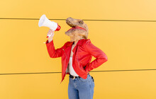 Young Woman Wearing Dinosaur Mask Talking Through Megaphone In Front Of Yellow Wall