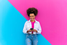 Smiling Young Woman With Mobile Phone Standing In Front Of Pink And Blue Wall