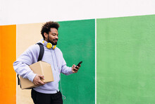 Delivery Man Holding Box Using Smart Phone In Front Of Colorful Wall