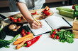 © Tijana - Close Up Photo of Woman Hand Writing Recipe in a Empty Notebook at Kitchen Desk with Many Vegetables
