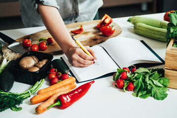 Close Up Photo of Woman Hand Writing Recipe in a Empty Notebook at Kitchen Desk with Many Vegetables 