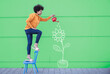 © VISTA by Westend61 - Woman standing on chair watering flowers painted on wall