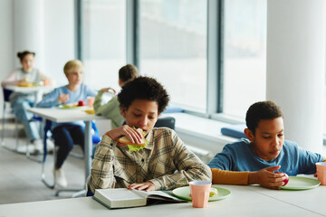 Wall Mural - Portrait of schoolchildren at lunch break, focus on young african American girl eating sandwich and reading book, copy space