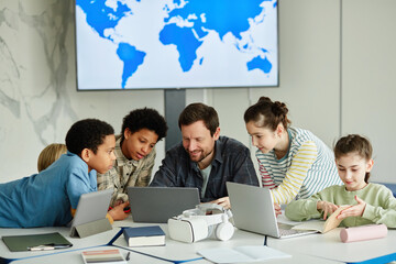 Wall Mural - Front view portrait of smiling male teacher with diverse group of children using laptop together in modern school classroom