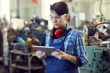 Wall Mural - Serious young brutal woman in protective goggles and blue overall standing in workshop and reading technical specification on tablet