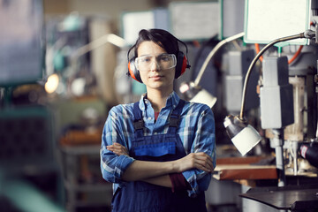 Wall Mural - Portrait of content confident skilled young woman in ear protectors and safety goggle working at factory with manual lathes