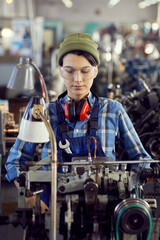 Wall Mural - Serious concentrated young female mechanic in protective goggles and hipster hat standing in industrial workshop and repairing machine