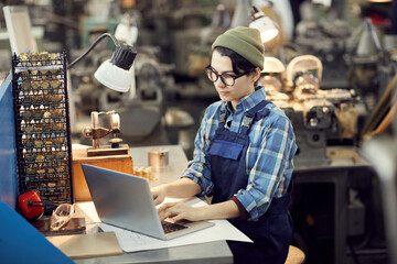 Wall Mural - Concentrated hipster girl in beanie hat sitting at table in watch factory shop and using laptop while viewing files of technical project