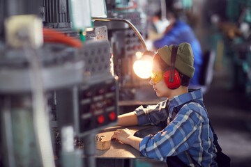 Wall Mural - Side view of young female processing worker in ear protectors and yellow goggles sitting at desk and using manual machine for polishing metal details