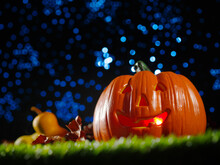 Against The Background Of A Blue Starry Sky, An Orange Pumpkin With A Smile, Illuminated From The Inside, Pears, Apples, Cones On Green Grass. Halloween Autumn Holiday Celebration.