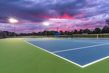 Evening Photo Of Outdoor Blue Tennis Courts With Pickleball Lines With Lights Turned On.	