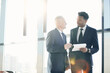 © Mediaphotos - Content multi-ethnic business experts in suits standing at window in office and looking through papers while thinking of company strategy