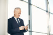 © Mediaphotos - Serious busy gray-haired businessman in dark suit leaning on wall in modern lobby with panoramic windows and making notes in smartphone