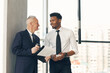 © Mediaphotos - Positive confident senior businessman with coffee cup holding clipboard with papers and discussing project implementation with young black PR manager