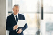 © Mediaphotos - Serious senior manager with gray hair standing in lobby with panoramic window and examining papers