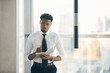 © Mediaphotos - Serious thoughtful young black manager in white shirt and tie standing at window and writing down ideas in diary