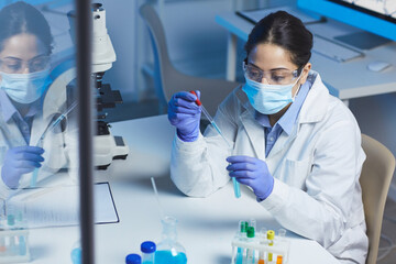 serious young indian female chemical researcher in protective workwear sitting at desk in laboratory