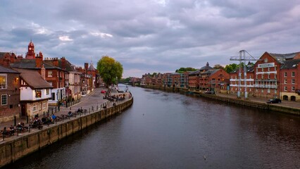 Canvas Print - York, UK. People walk by the embankment area of the Ouse river in York, UK. Time-lapse of heavy sunset clouds and touristic boats. Cloudy evening