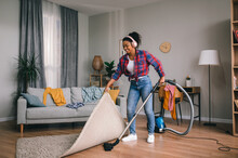 Happy young african american woman in headphones vacuuming with vacuum cleaner in living room