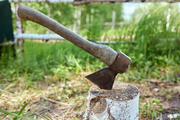 A large old ax sticks out of a wooden stump chopping block against the background of a green clearing of clover on a summer sunny day.